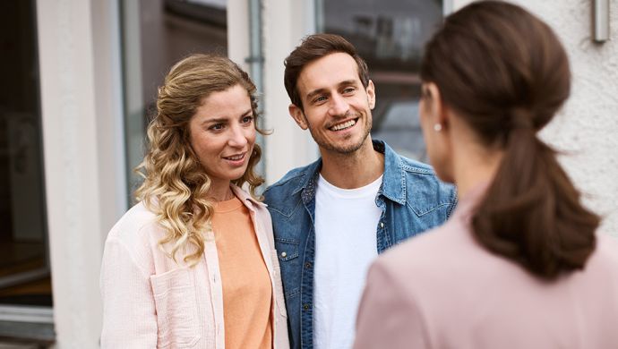Junges Paar im Gespräch mit einer Maklerin vor einem Gebäude. Freundliche Interaktion, möglicherweise bei einer Immobilienbesichtigung oder Beratungsgespräch. Symbol für Hauskauf, Immobilienberatung und Kundenservice.